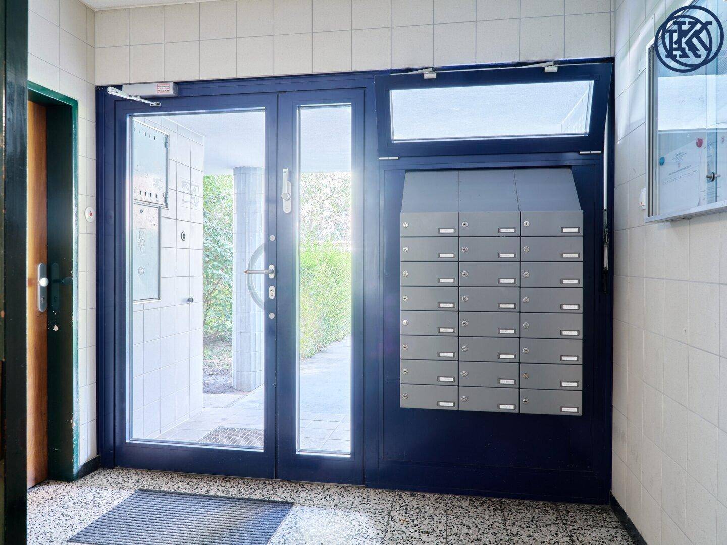 Eingangsbereich eines Mehrfamilienhauses mit Briefkästen und Glastür.