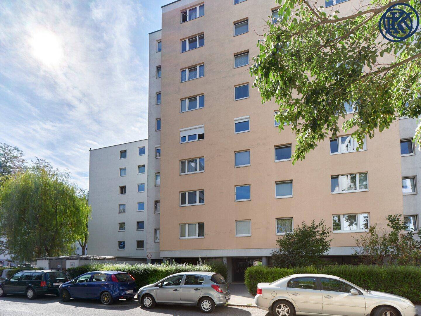 Ansicht eines mehrstöckigen Wohnhauses mit beiger Fassade und geparkten Autos auf der Straße.