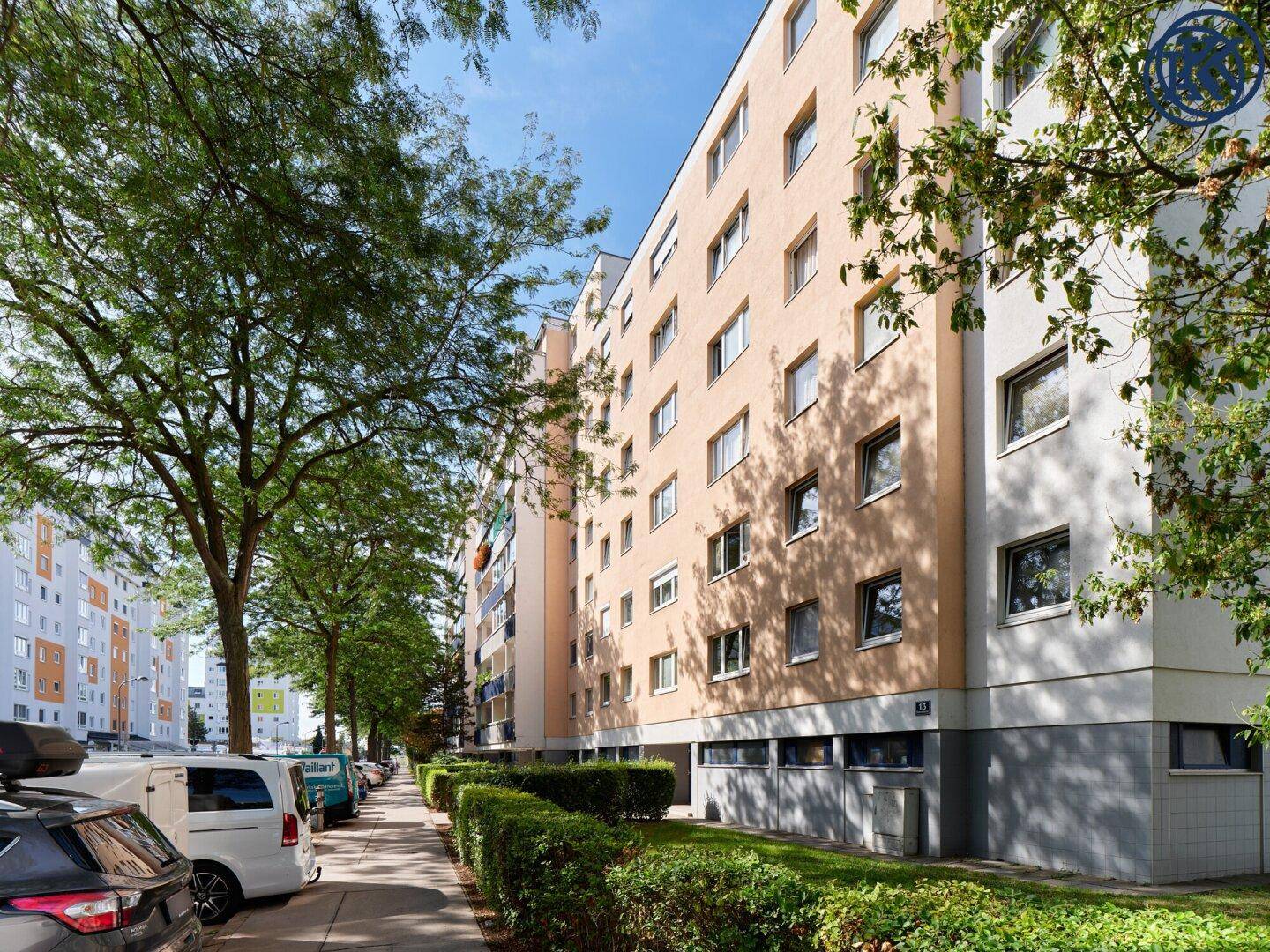 Mehrstöckiges Wohngebäude mit beiger Fassade und vielen Fenstern, umgeben von Bäumen und Parkplätzen.
