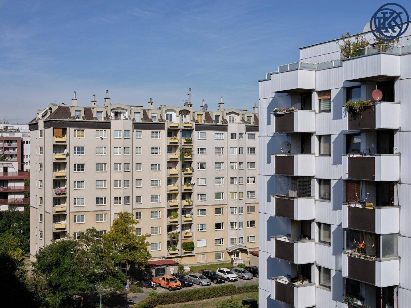 Mehrere Wohngebäude mit Balkonen und umliegenden Bäumen sowie geparkten Autos unter blauem Himmel.