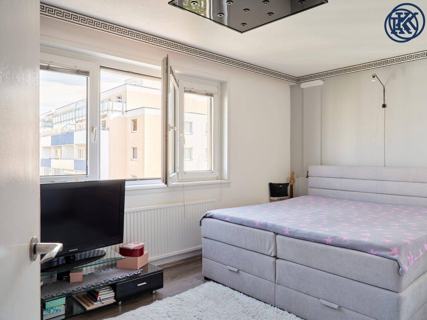 Helles Schlafzimmer mit Bett, Fernseher und großen Fenstern, dekorative Deckenleiste.