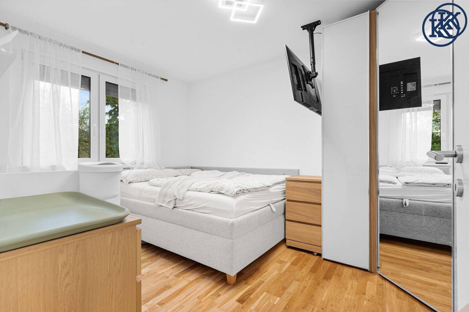 Helles Schlafzimmer mit Doppelbett, Holzkommode und wandmontiertem Fernseher.