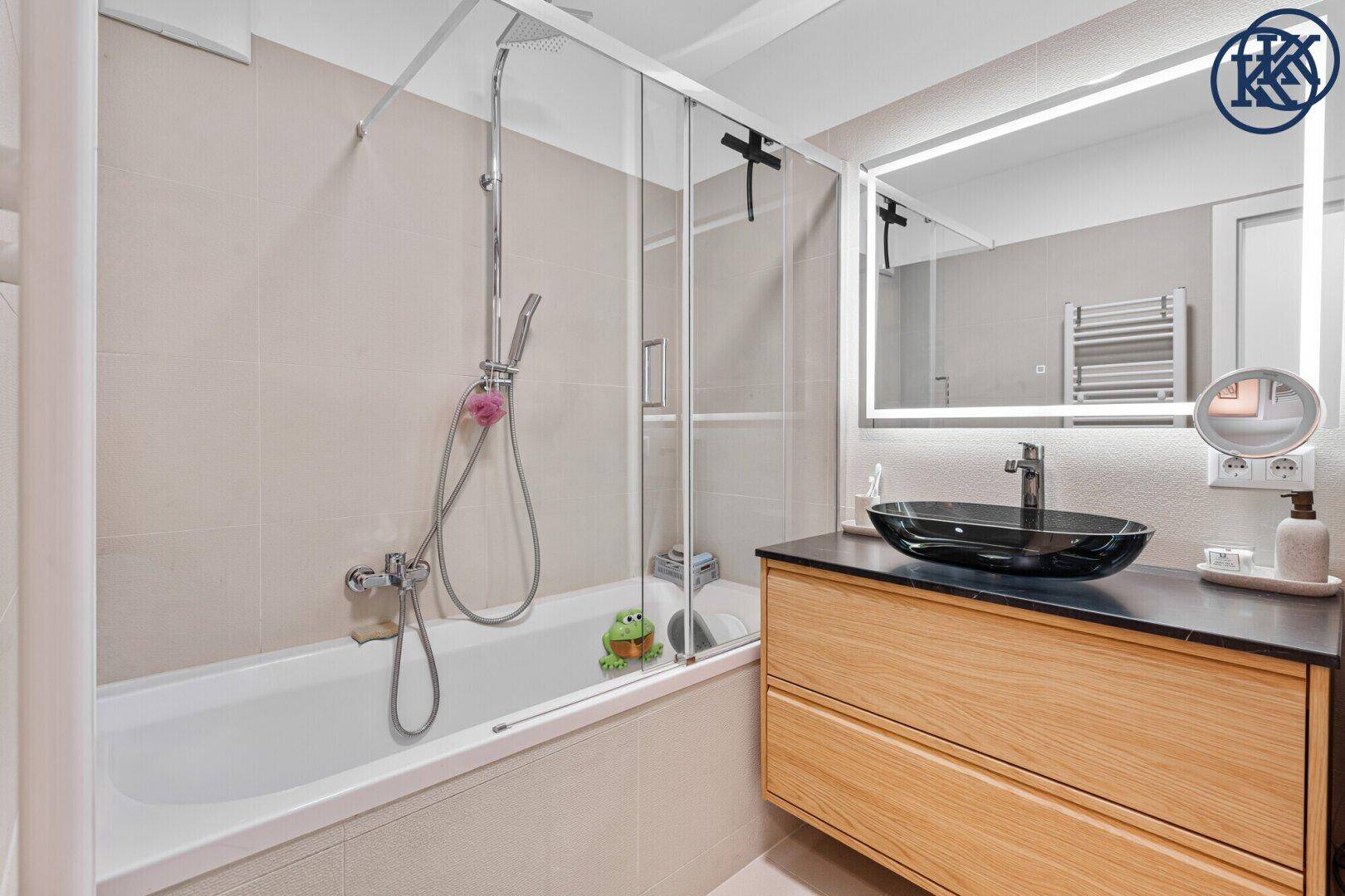 Helles Badezimmer mit Badewanne, Glasduschwand und modernem Waschtisch mit beleuchtetem Spiegel.