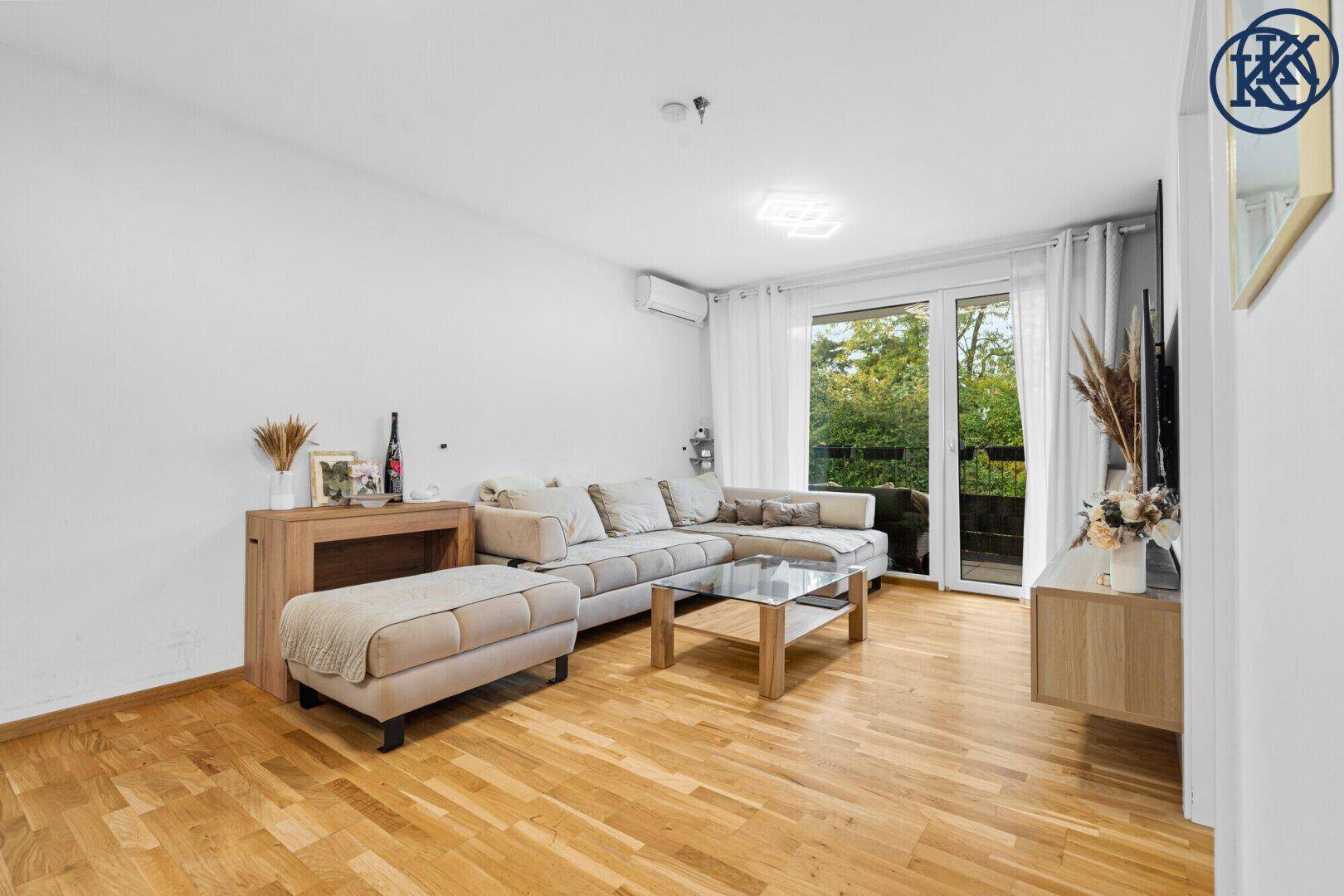 Geräumiges Wohnzimmer mit hellem Holzboden, Ecksofa und großem Fenster zum Balkon.