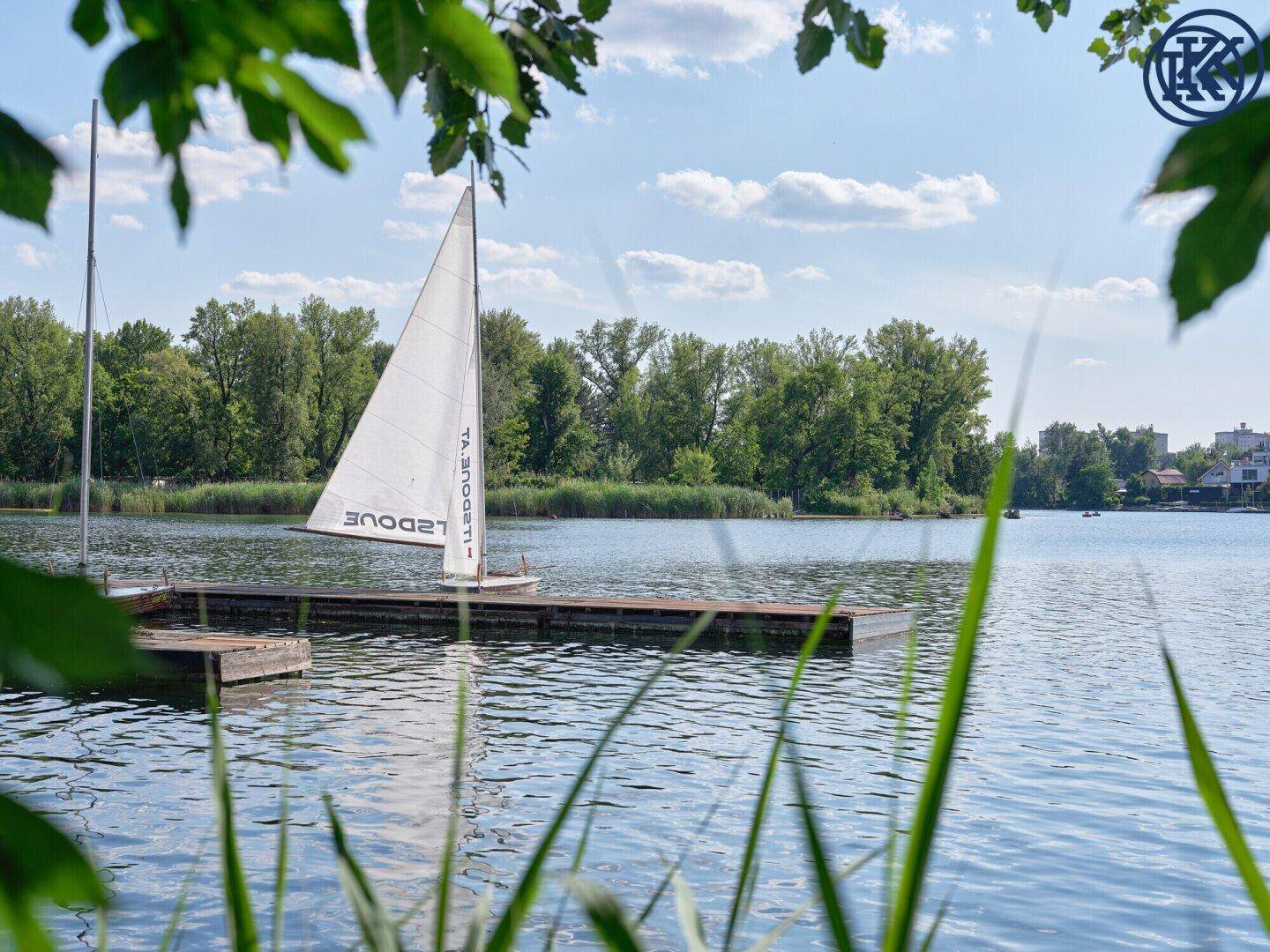 Idyllischer Seeblick mit einem kleinen Segelboot an einem Steg unter blauem Himmel.