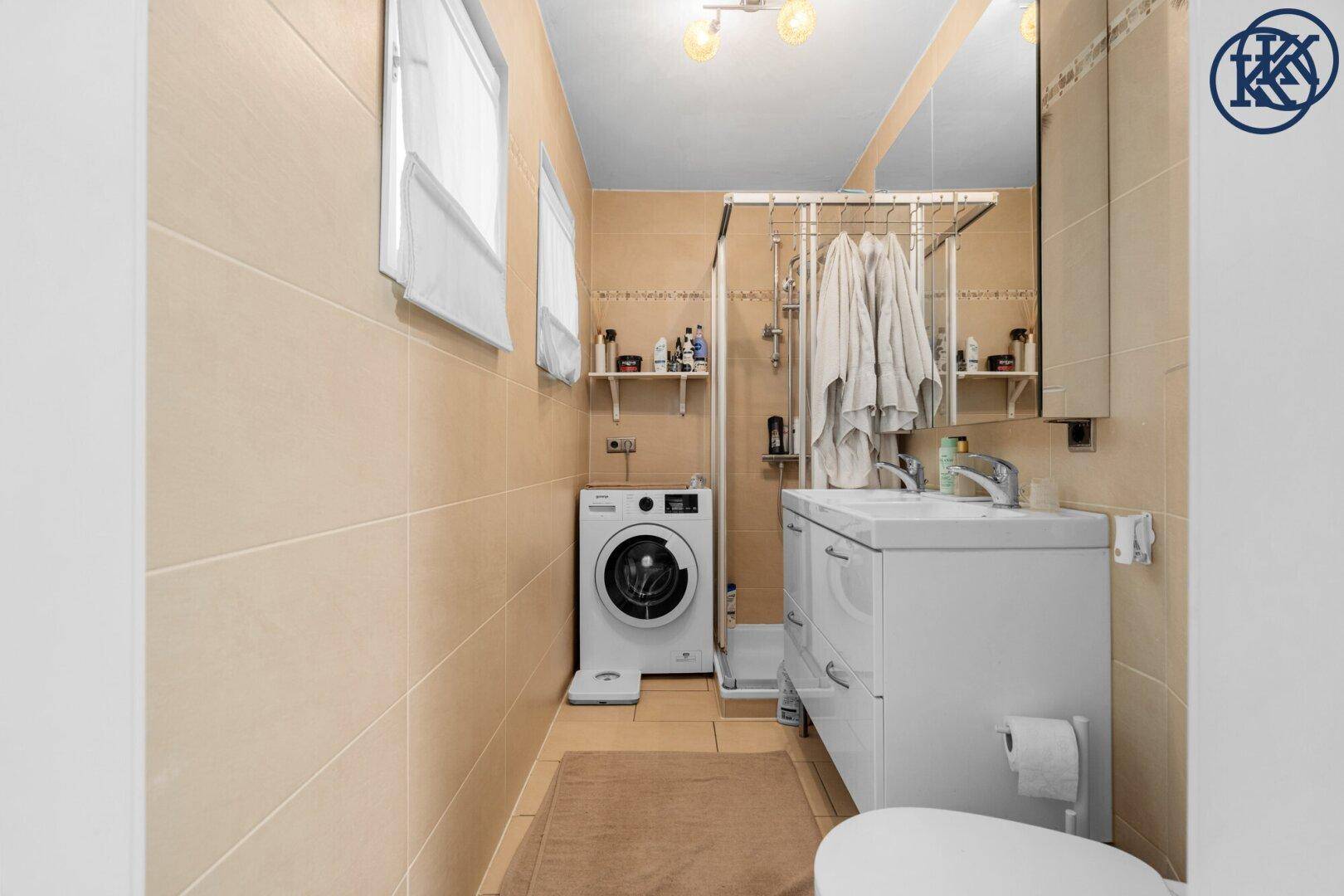 Badezimmer mit Duschkabine, Waschbecken und Waschmaschine, komplett mit beigen Fliesen ausgestattet.