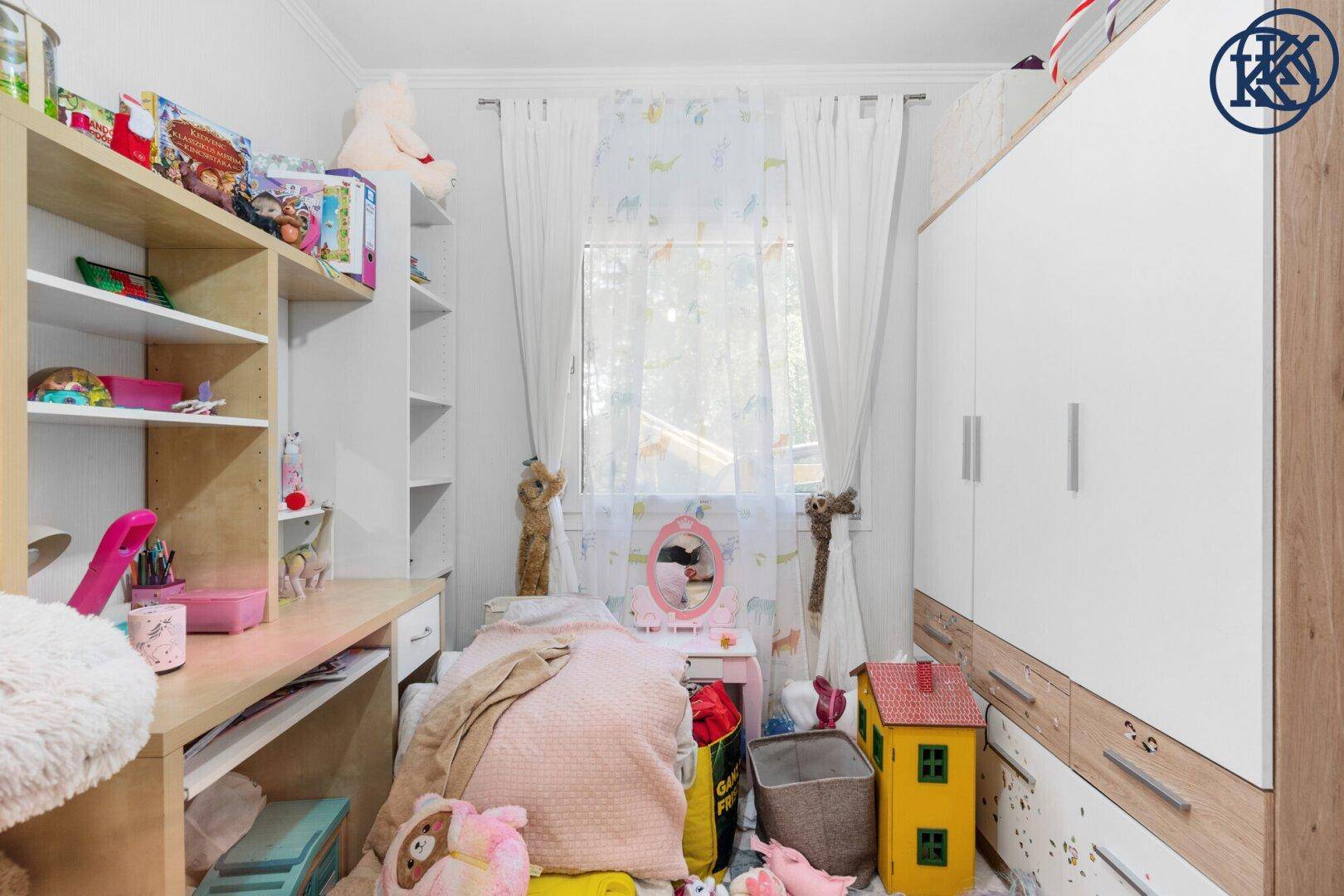 Helles Kinderzimmer mit Schreibtisch, Regalen und Blick ins Grüne durch das Fenster.