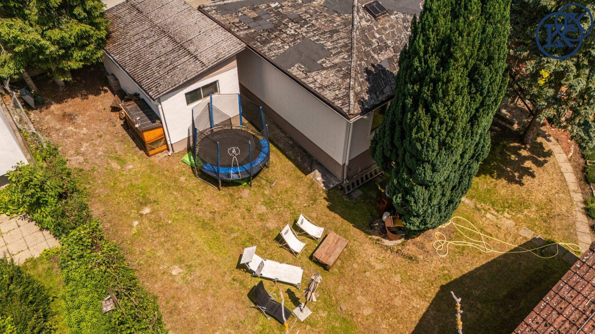 Gartenbereich mit Trampolin, Liegestühlen und einem großen Baum neben dem Haus.