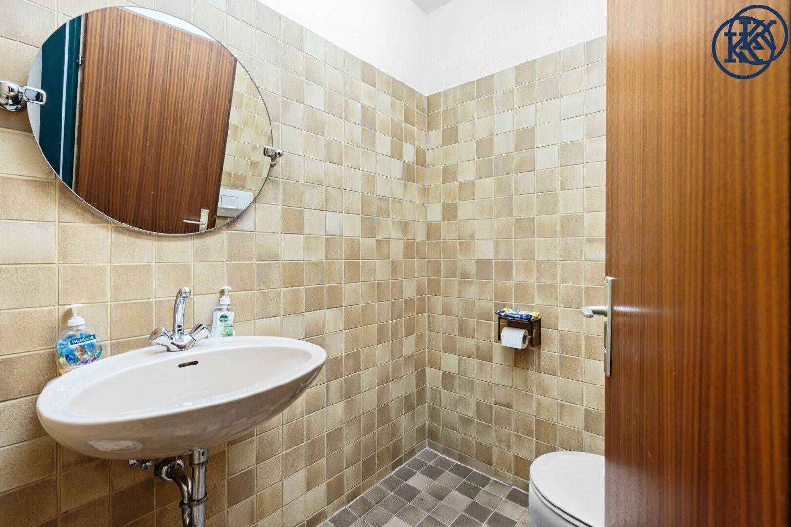 Badezimmer mit beigefarbenen Fliesen, Waschbecken und Toilette, ein runder Spiegel hängt an der Wand.