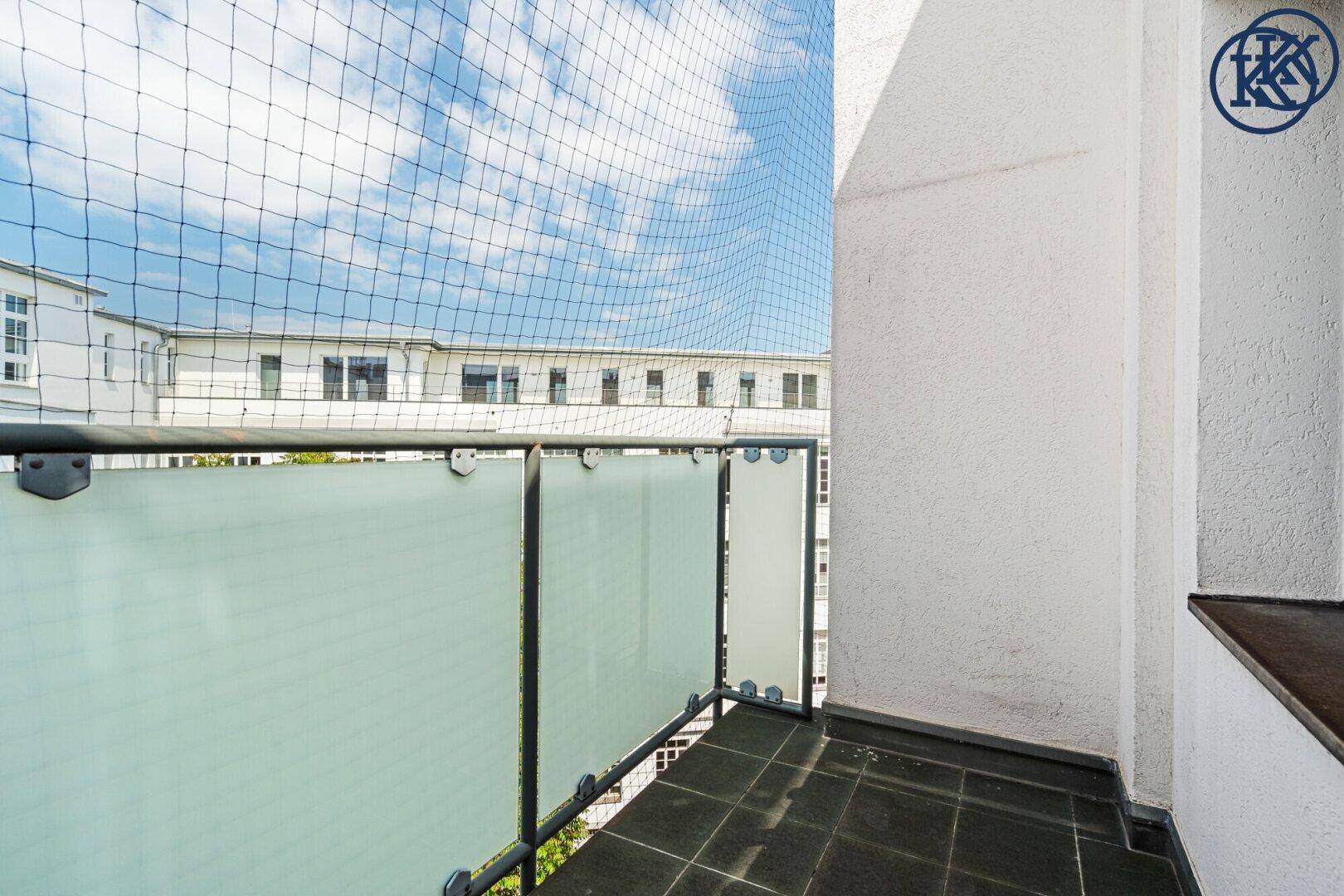 Balkon mit Glasbrüstung und Schutznetz, Blick auf umliegende Gebäude und den Himmel.