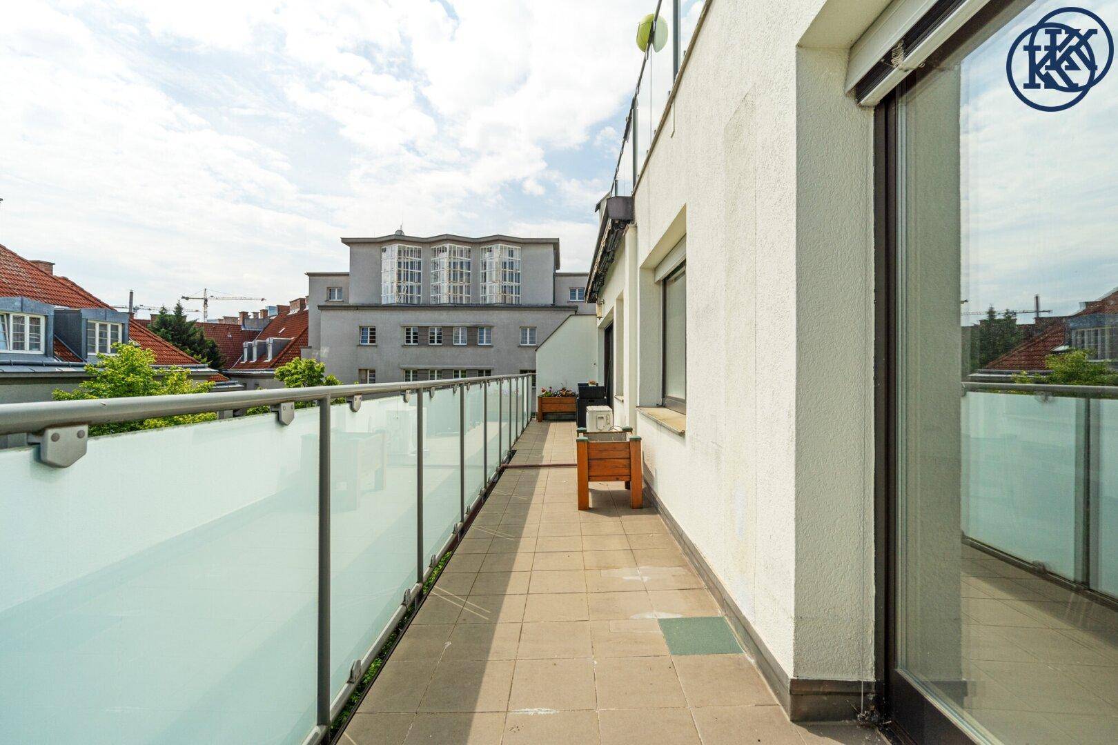 Lange Terrasse mit Fliesenboden und Glasbrüstung, Blick auf die Dächer der Nachbargebäude.