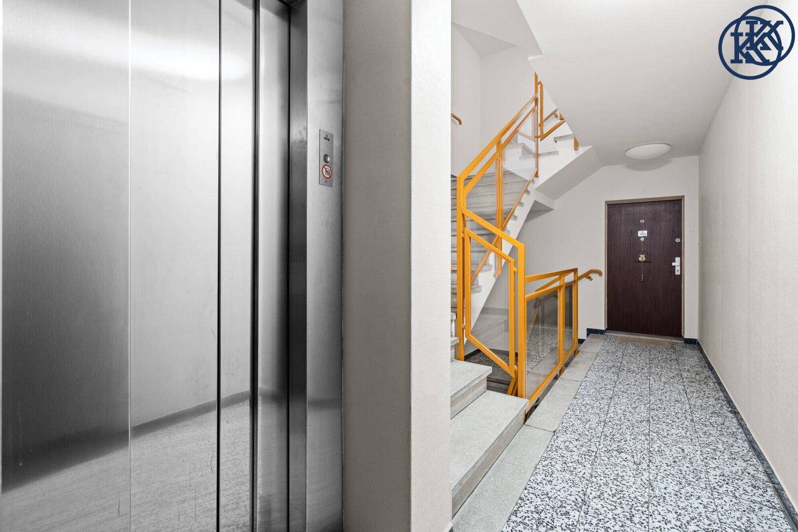 Treppenhaus mit Aufzug und gelbem Geländer, Terrazzoboden und Holztür zum Apartment.