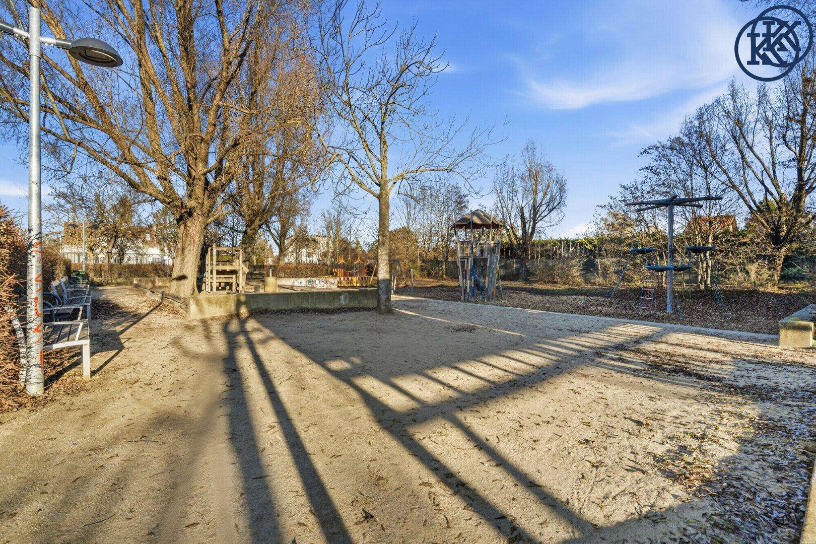 Großer Spielplatz mit Sandfläche, Bäumen und verschiedenen Spielgeräten für Kinder.