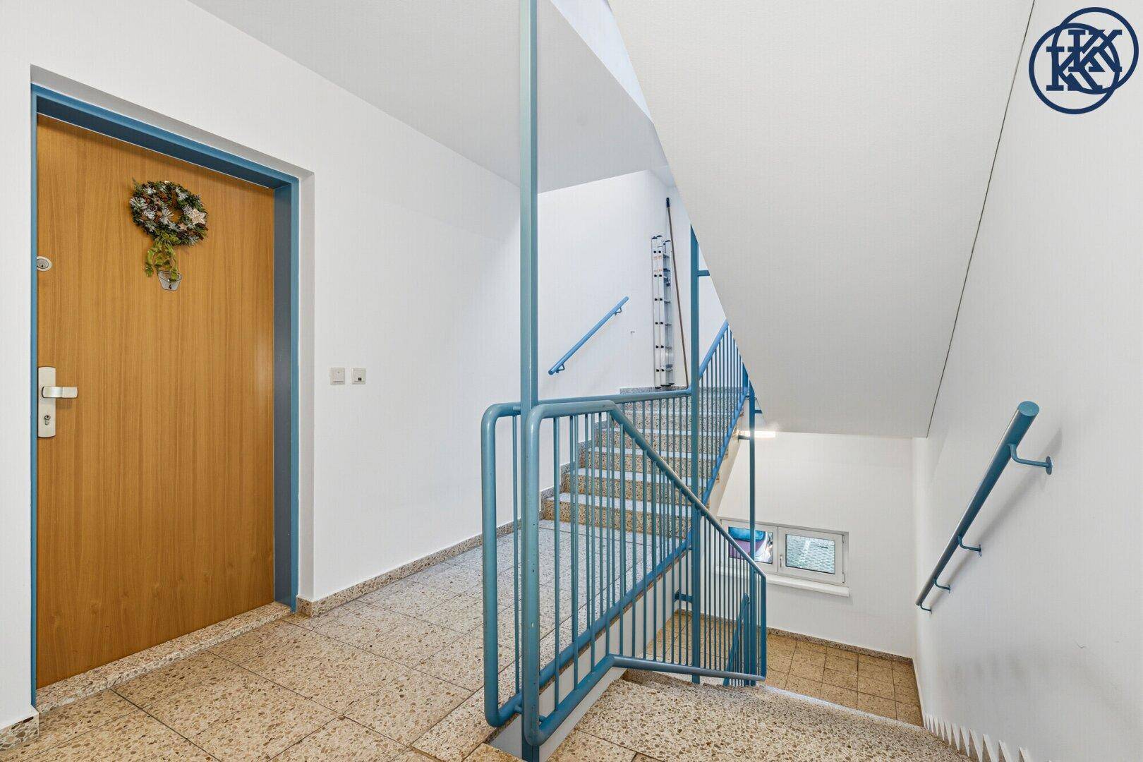 Helles Treppenhaus mit blauen Geländern und robustem Terrazzoboden in einem Mehrfamilienhaus.