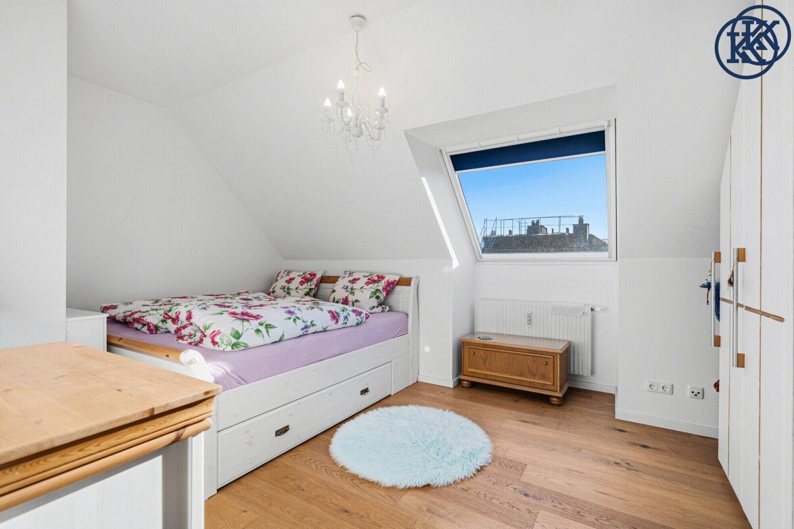 Schlafzimmer mit Holzboden, Bett und Dachschräge, beleuchtet durch ein großes Dachfenster.
