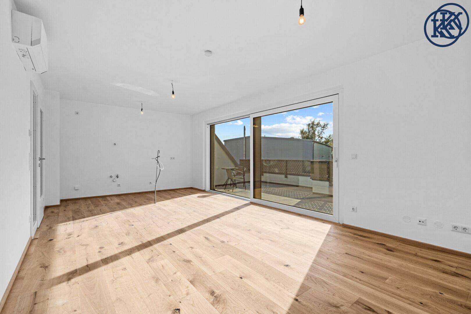 Leerer offener Wohnbereich mit Holzboden und großer Glasschiebetür zur Terrasse, sehr hell.