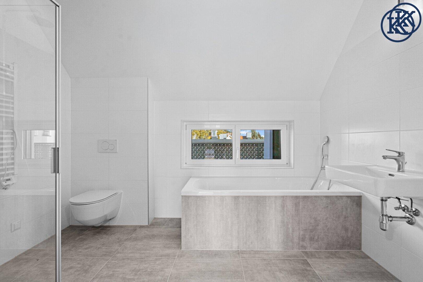 Modernes Badezimmer mit Badewanne, WC, Waschbecken und Fenster, hell gefliest in Weiß und Grau.