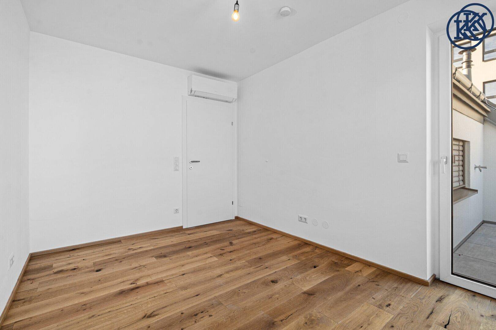 Leeres Zimmer mit Holzboden, Klimaanlage und Zugang zu einem Außenbereich, ideal als Schlafzimmer.
