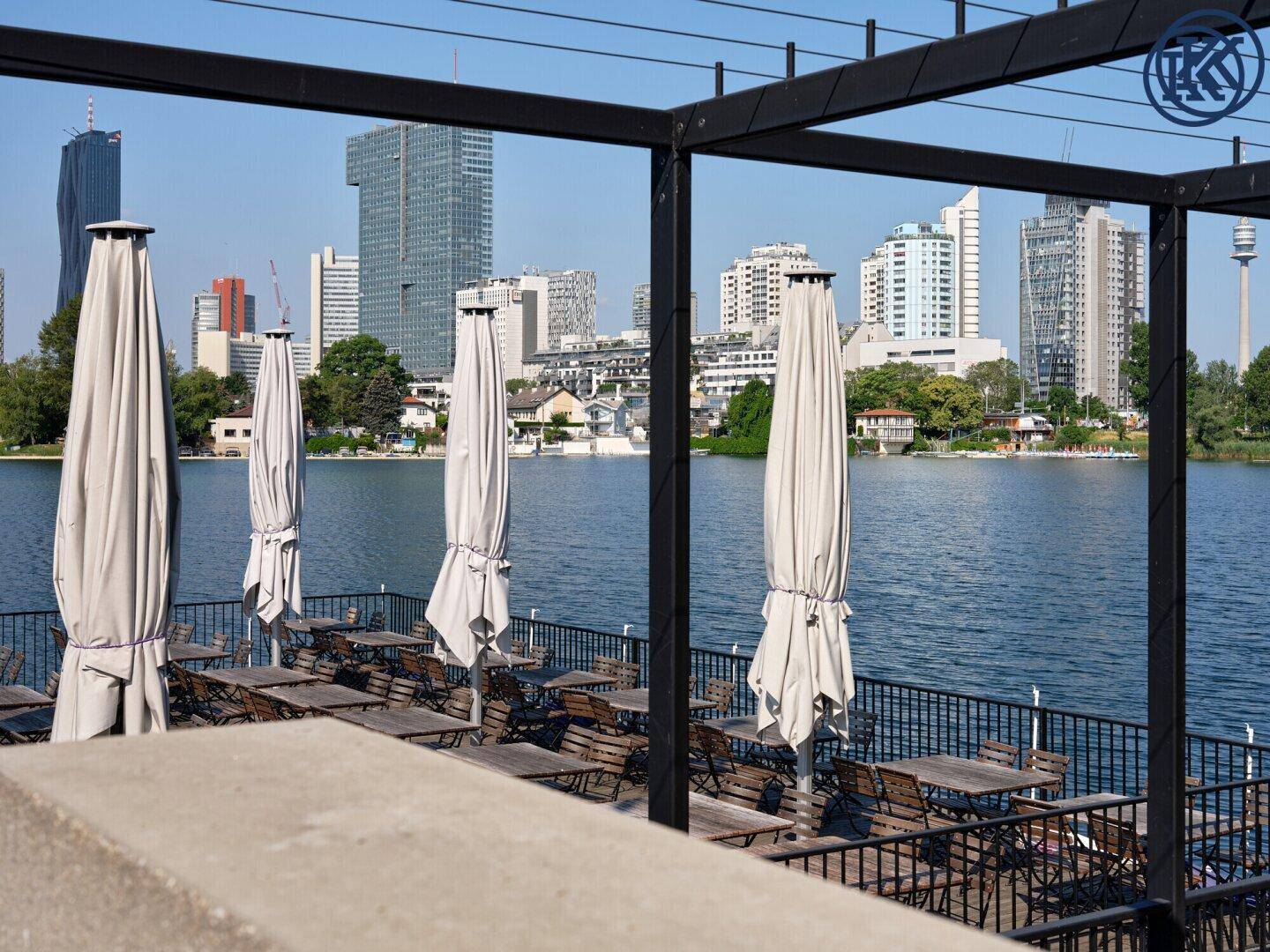 Restaurantterrasse am Seeufer mit Tischen, Sonnenschirmen und Stadtblick im Hintergrund.