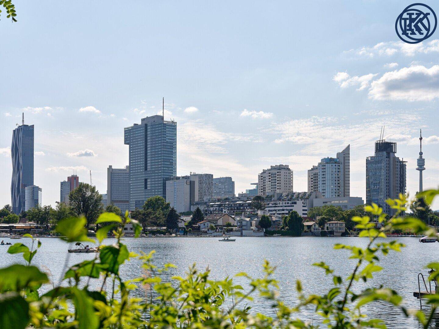 Blick auf die Wiener Skyline und den Kaiserwassersee mit grüner Ufervegetation im Vordergrund.