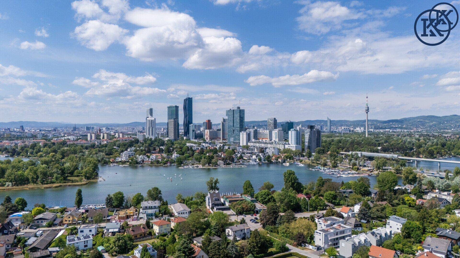 Panoramablick auf eine Stadt mit See, Hochhäusern und grünen Ufern unter blauem Himmel.