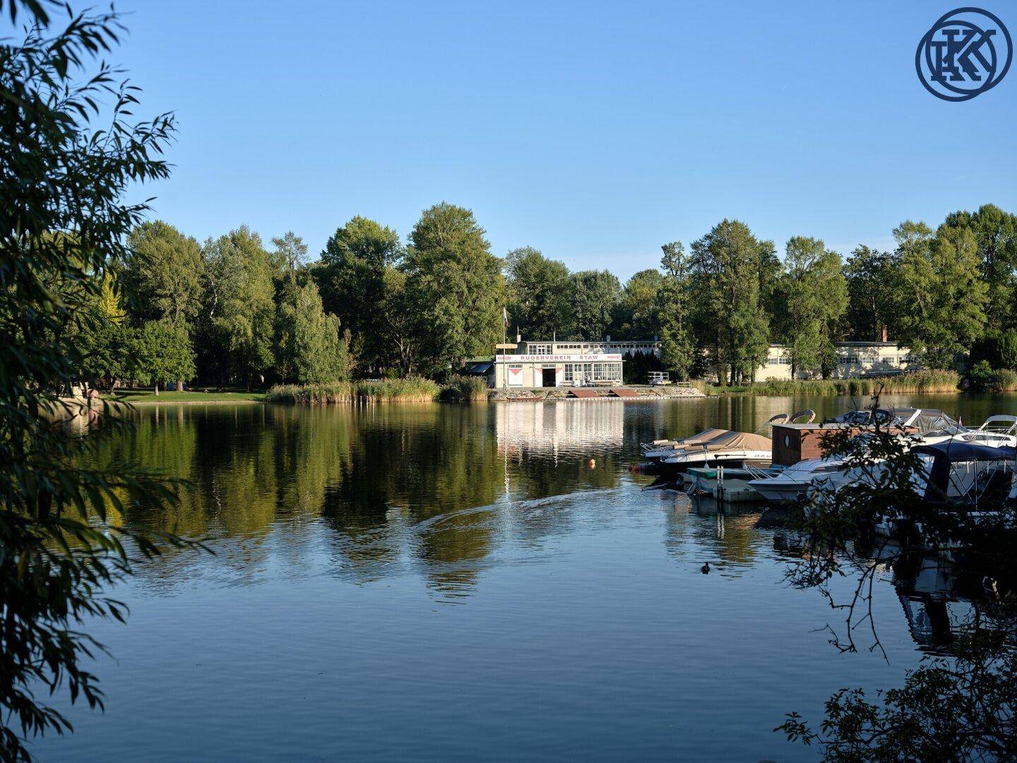 Idyllischer See mit festgemachten Booten und einem Gebäude am bewaldeten Ufer.