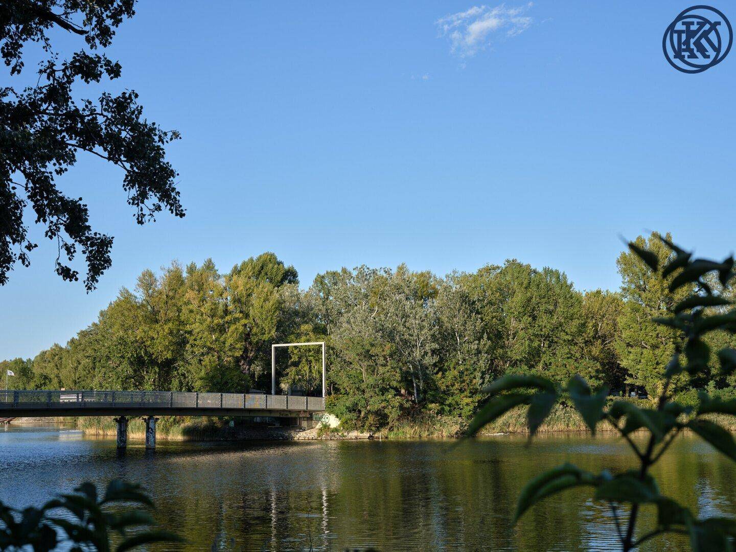 Brücke über einen Fluss, umgeben von üppigem Grün und Bäumen unter blauem Himmel.