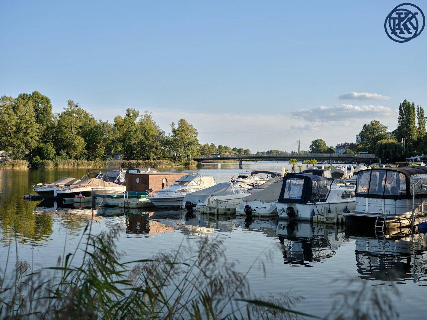 Boote liegen in einem Yachthafen am See, mit einer Brücke und Bäumen im Hintergrund.
