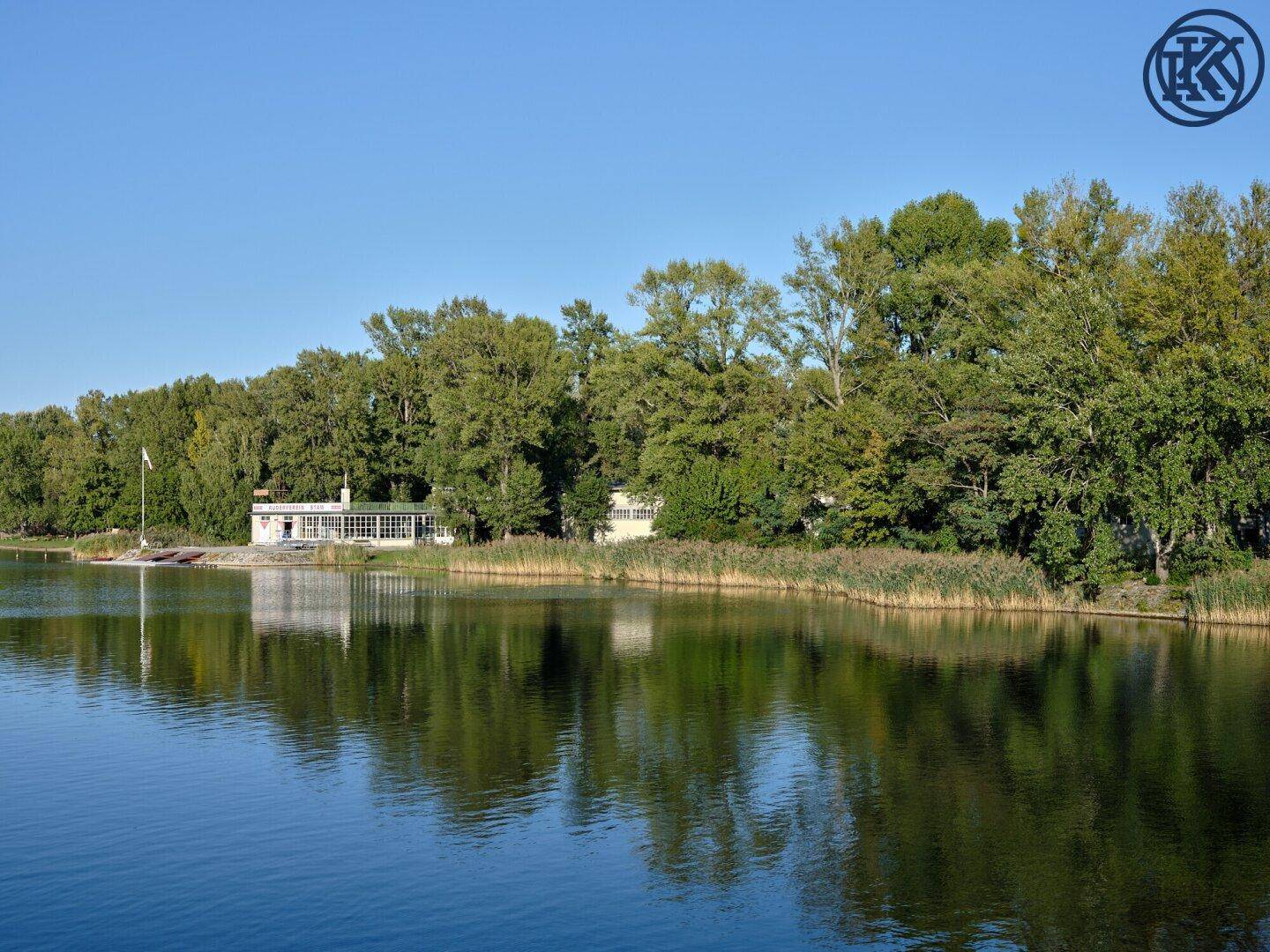 Ein weißes Gebäude mit großen Fenstern liegt idyllisch am Seeufer, umgeben von Schilf und Bäumen.