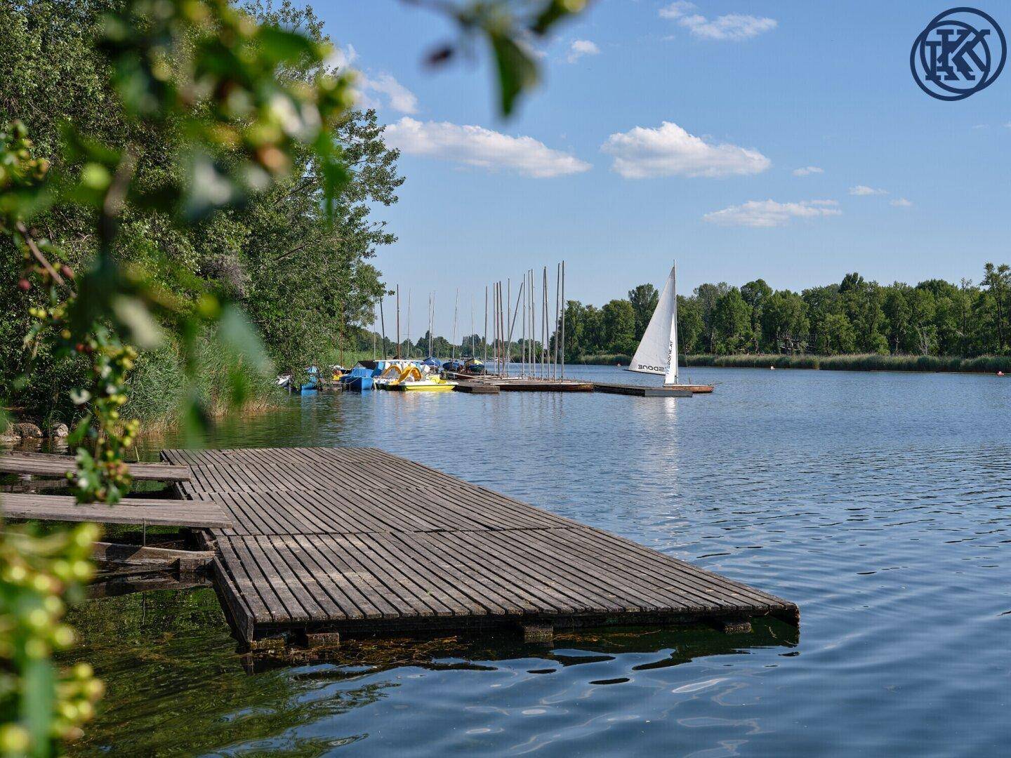 Holzsteg am See mit Segelbooten und einer malerischen Uferlandschaft im Hintergrund.