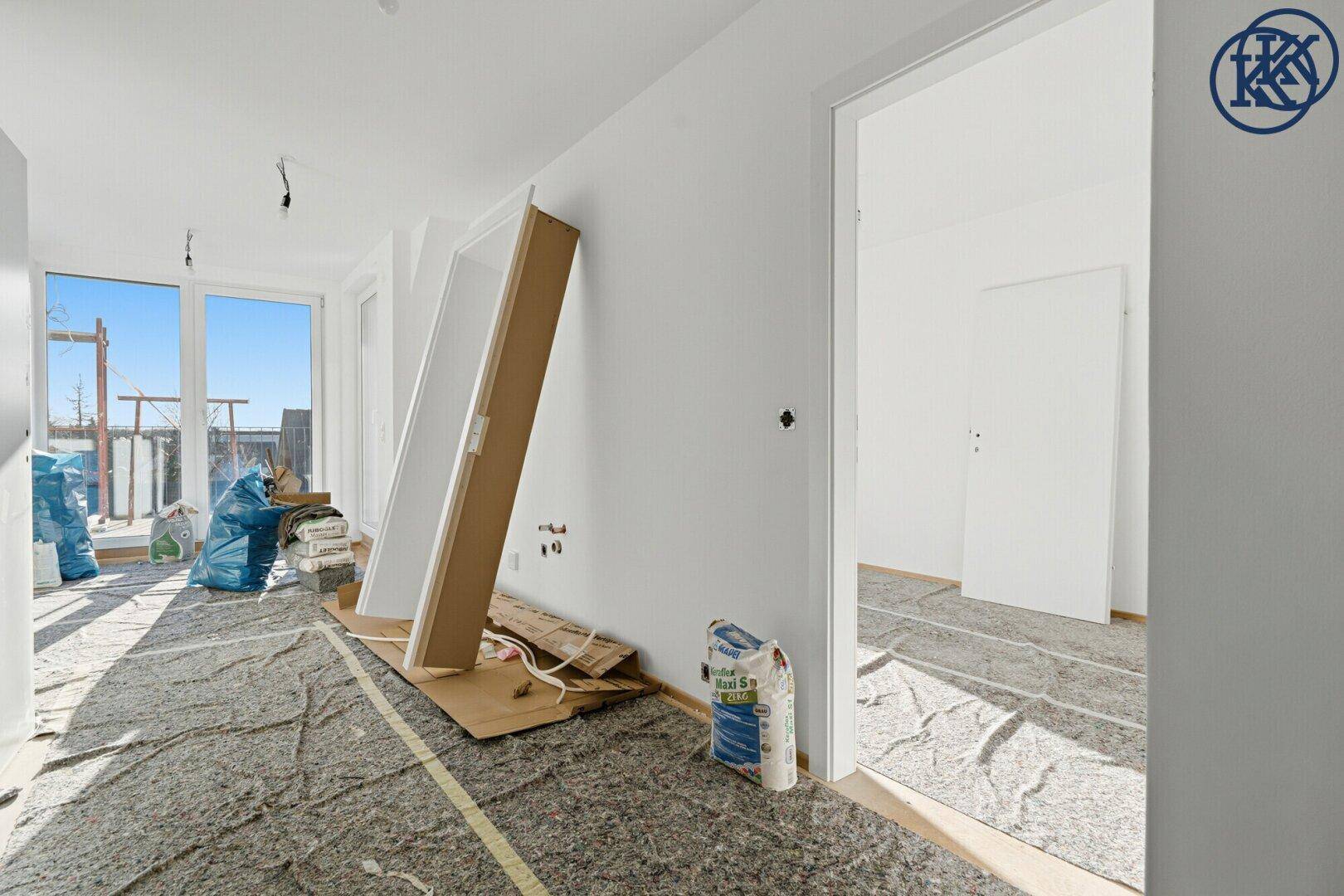 Helles Zimmer im Rohbau mit Blick nach draußen und Baumaterialien auf dem Boden.