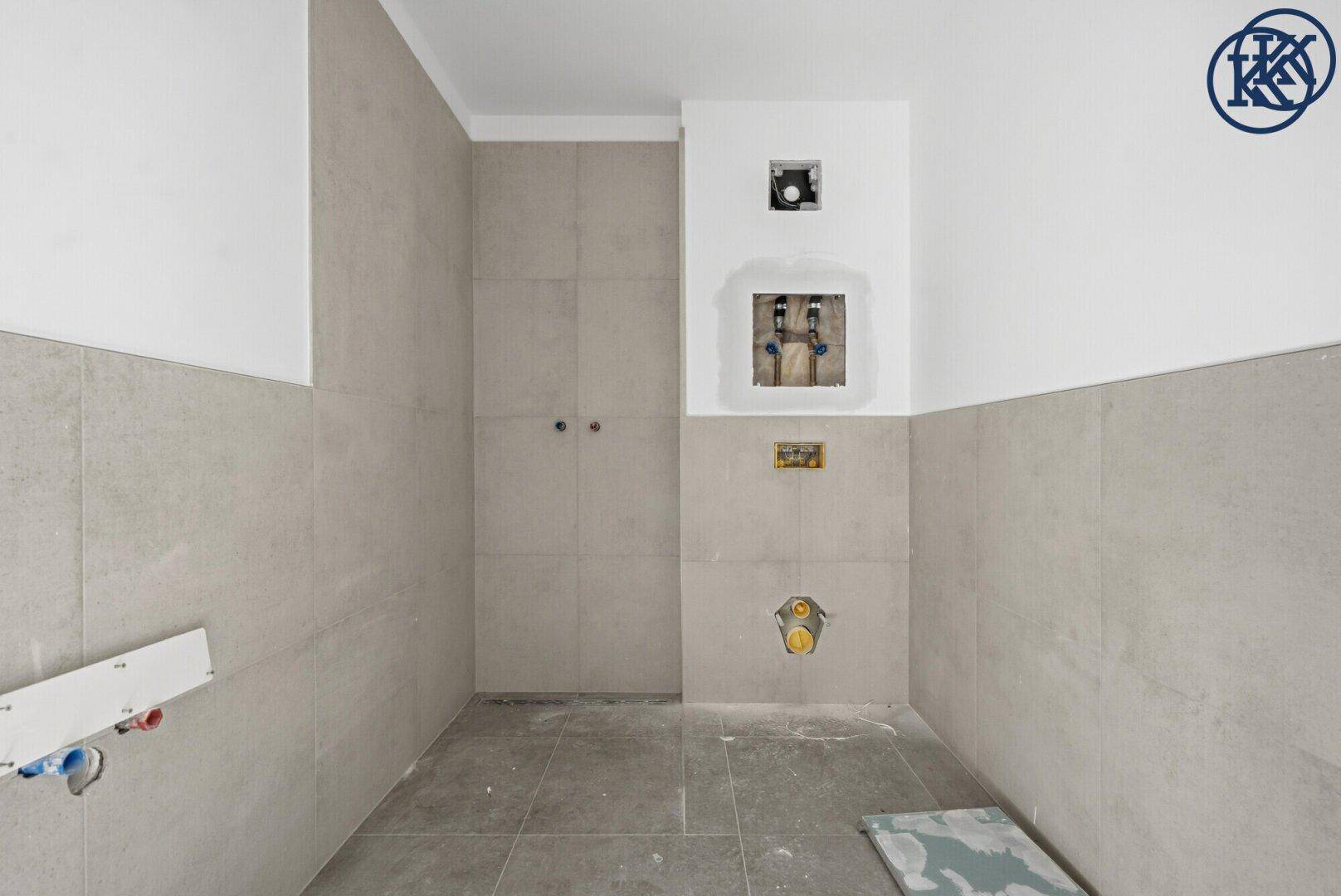 Badezimmer im Rohbau mit hellgrauen Fliesen und vorbereiteten Anschlüssen für Sanitäranlagen.