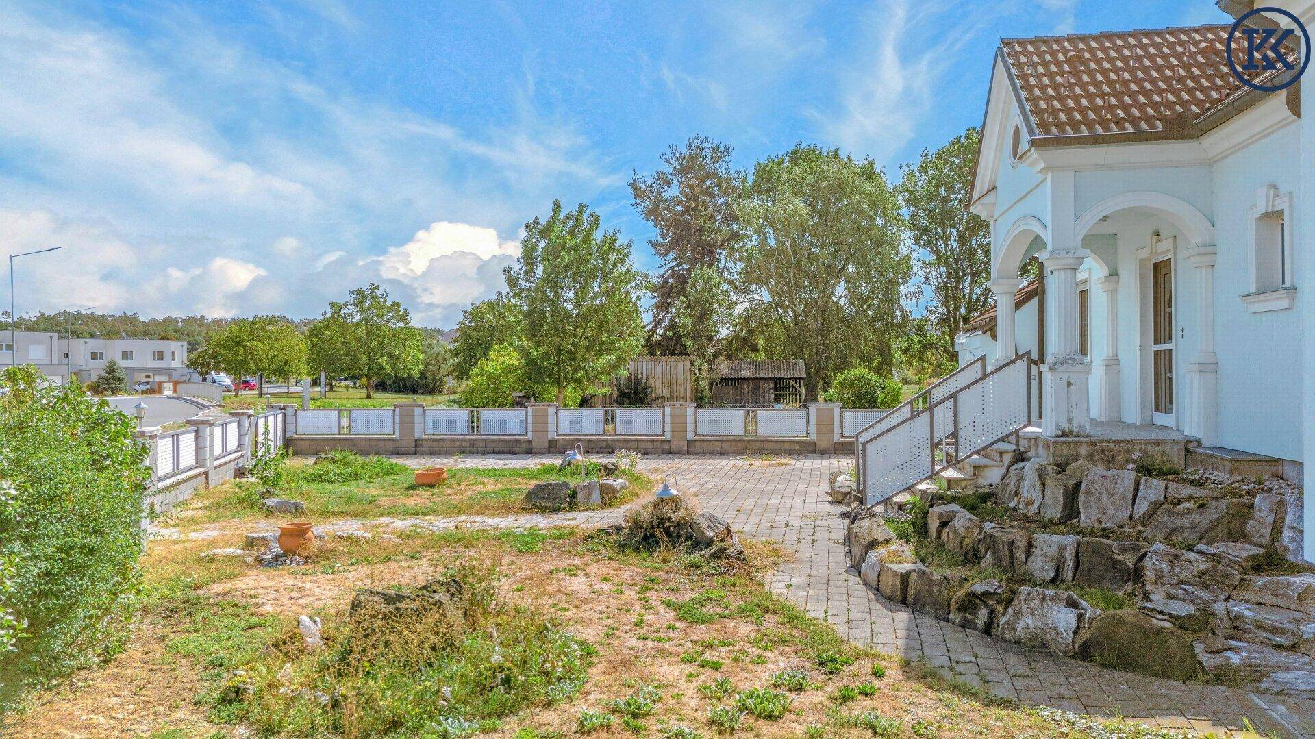 Blick vom Vorgarten über den Zaun auf die umliegende Landschaft und entfernte Gebäude.