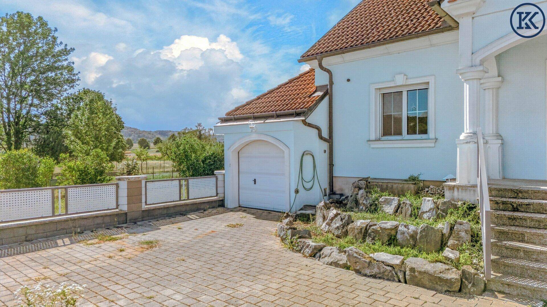 Integrierte Garage mit weißem Tor und gepflasterter Zufahrt, umgeben von Grünflächen.