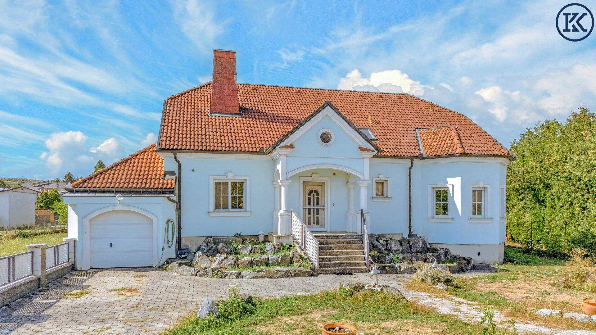 Großzügiges Einfamilienhaus mit hellblauer Fassade, rotem Ziegeldach und weitläufigem Vorgarten.