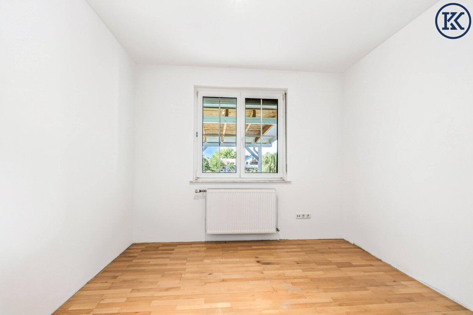 Freundliches Zimmer mit hellem Laminatboden, weißen Wänden und Blick ins Grüne.