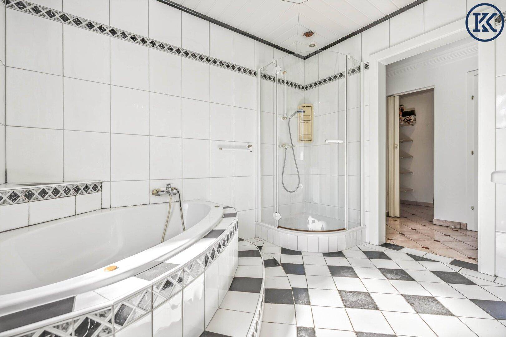 Badezimmer mit Duschkabine, Eckbadewanne und schwarz-weißem Fliesenboden, der zu einem weiteren Raum führt.