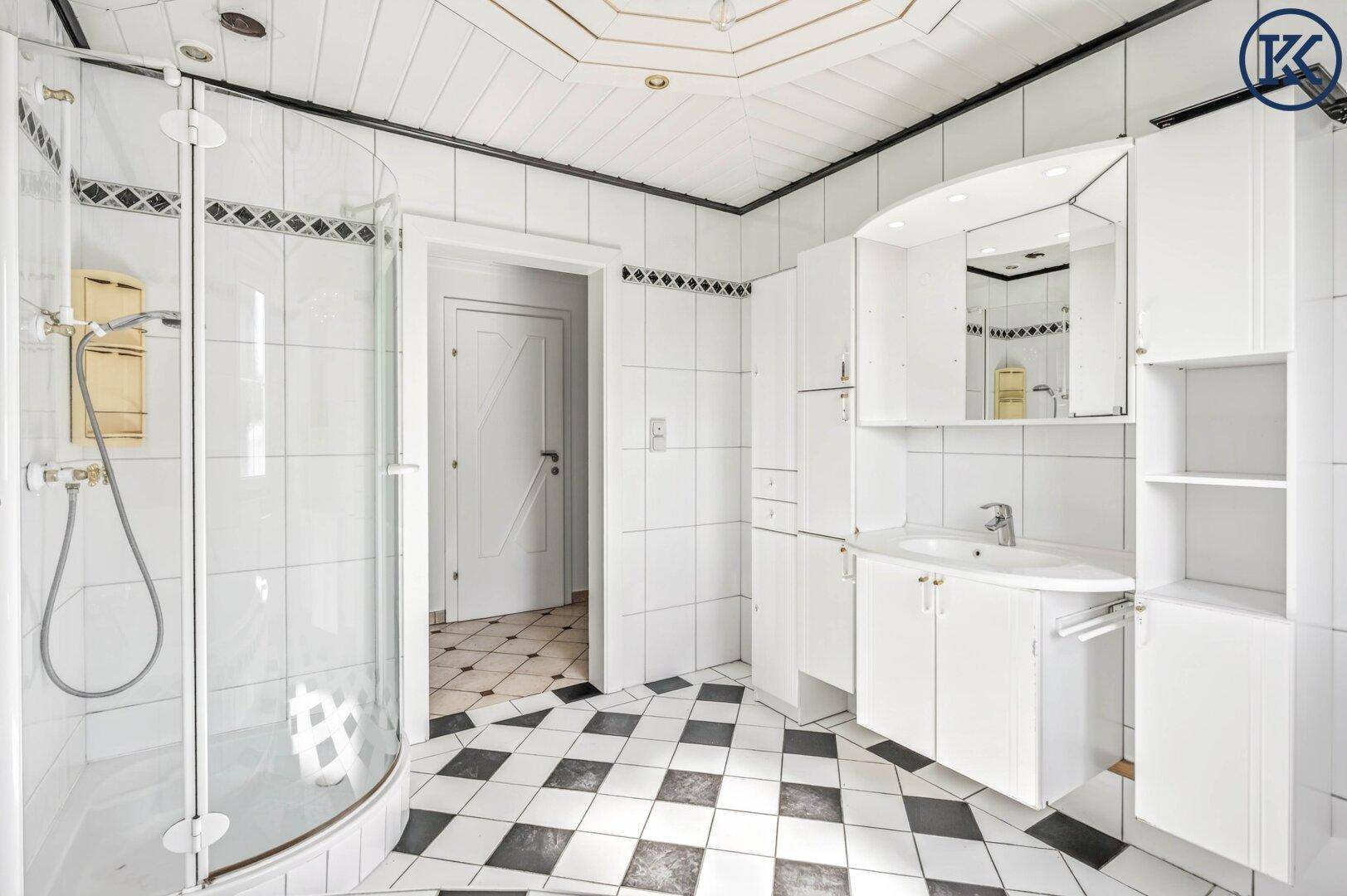 Badezimmer mit Duschkabine, weißem Waschtisch und schwarz-weißem Fliesenboden, der zum Flur führt.