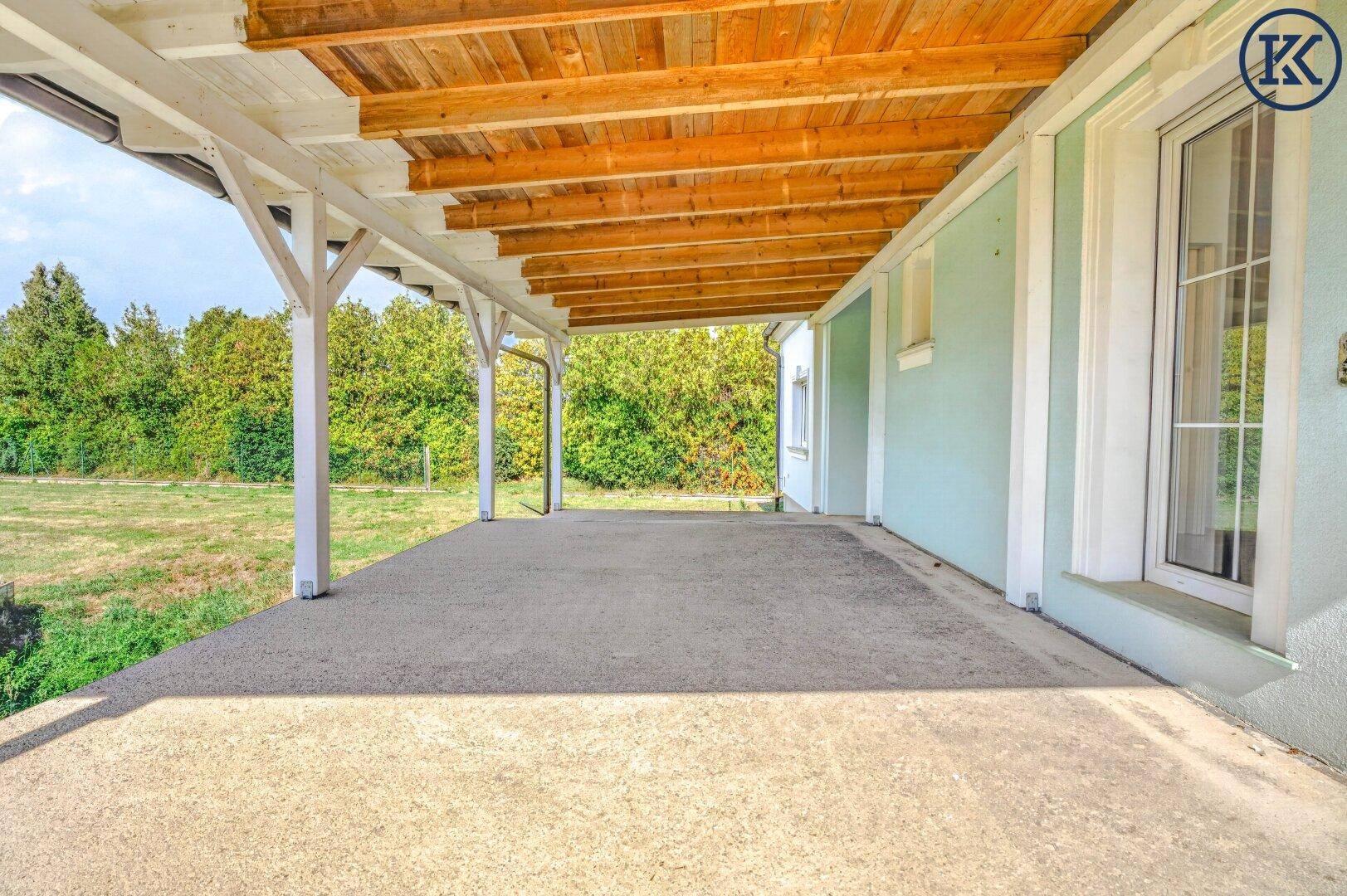 Überdachte Terrasse entlang der hellblauen Hausfassade mit Holzdach und Zugang zum Gartenbereich.