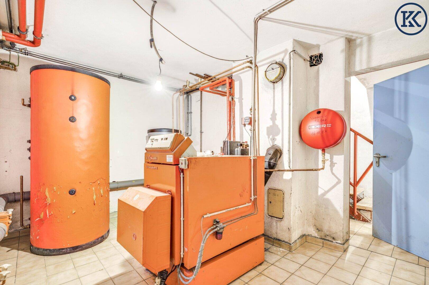 Heizungsraum im Keller mit großem orangefarbenem Boiler und sichtbaren Rohrleitungen.