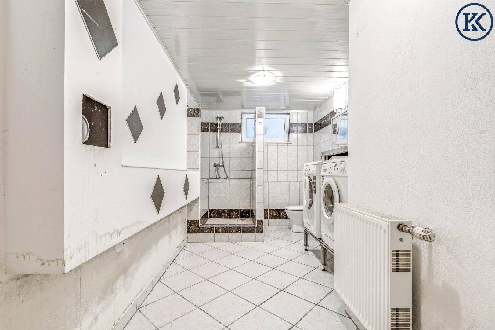 Kellerbad mit Dusche, WC, Waschmaschine und Heizkörper, ausgestattet mit Fliesen.