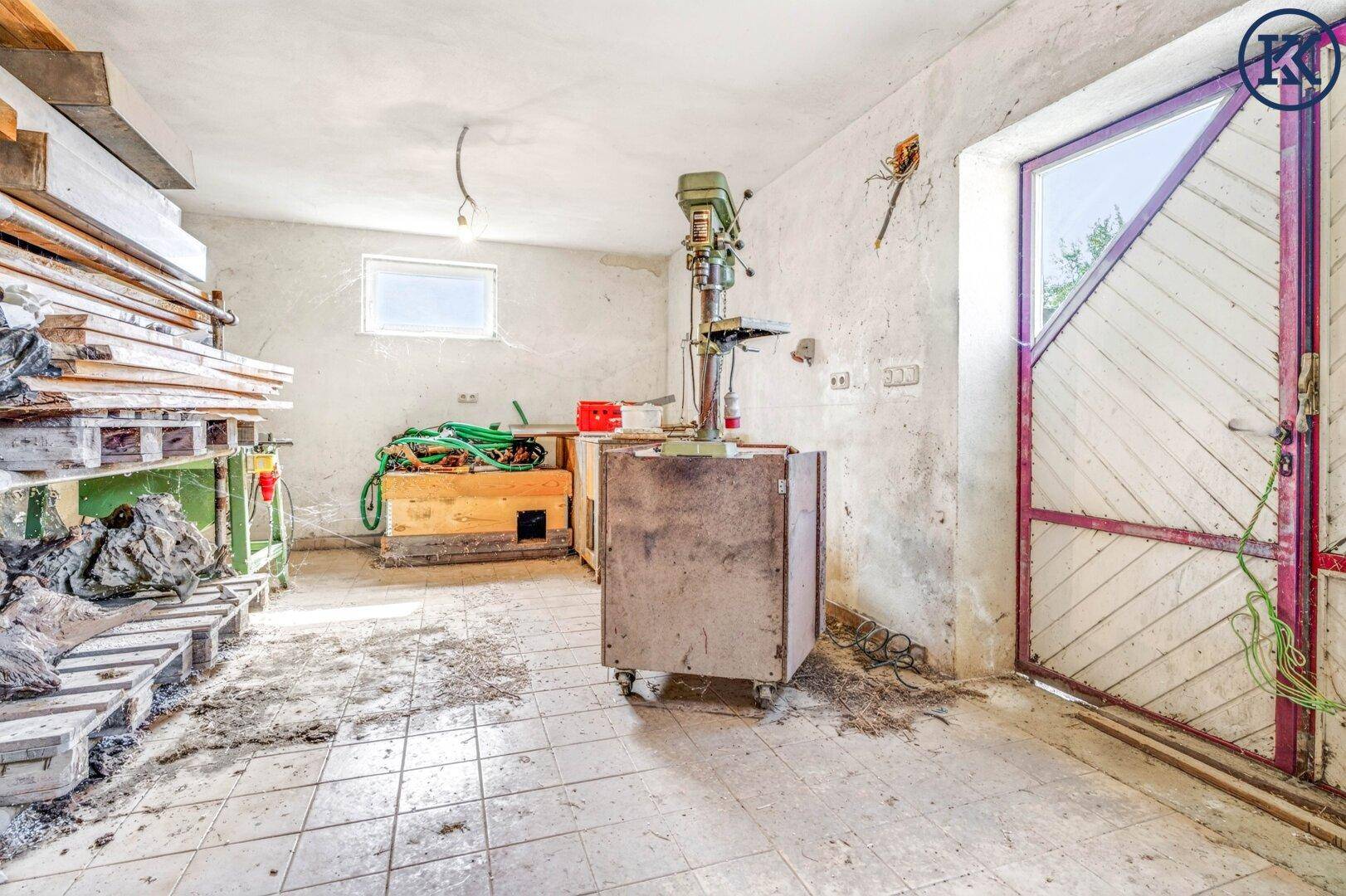Werkstattbereich im Keller mit Bohrmaschine, Holzpaletten, kleinem Fenster und rustikaler Holztür.