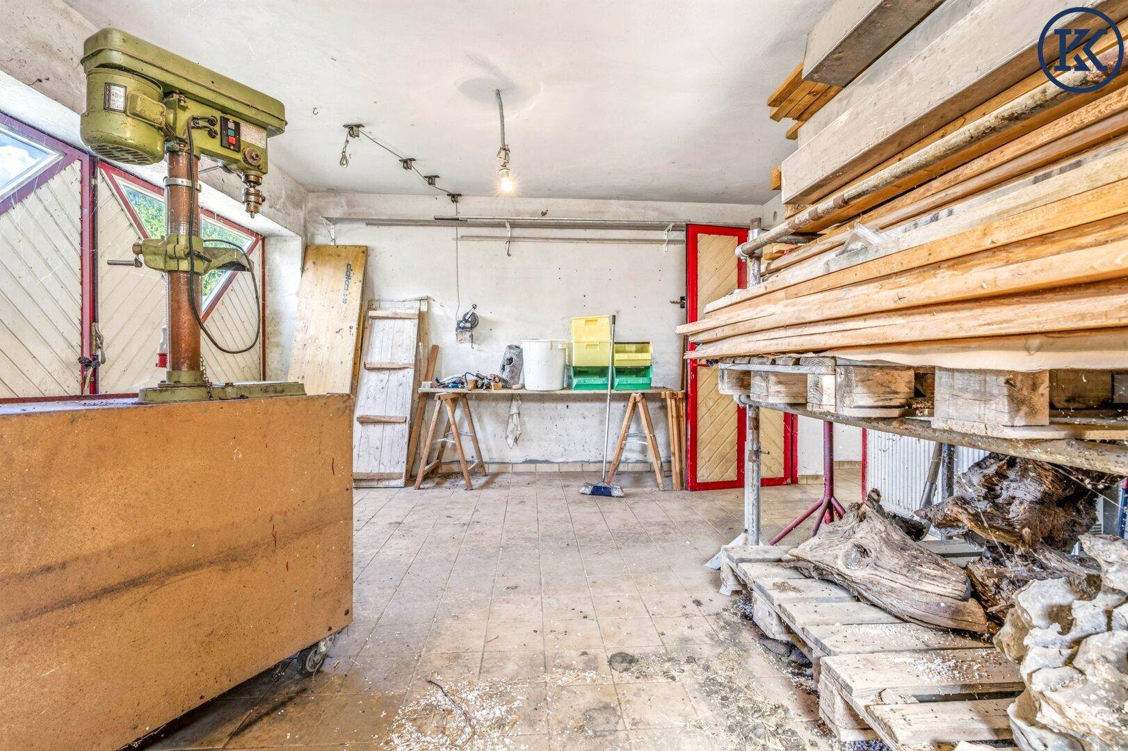 Werkstatt mit Bohrmaschine, Holzmaterialien und Arbeitsbereich auf gefliestem Boden, durch rustikale Türen zugänglich.