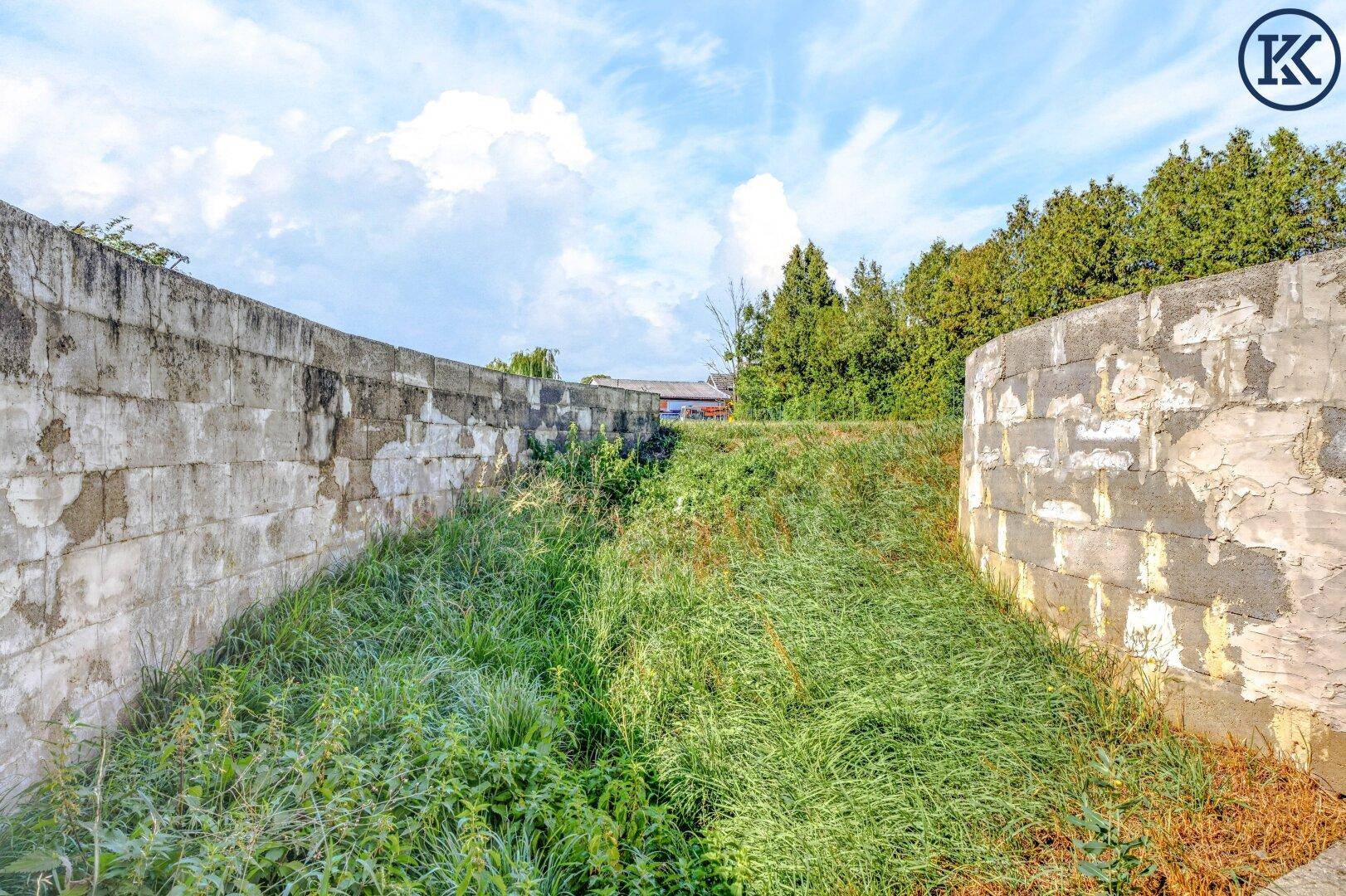 Außenbereich mit hohen Betonmauern und überwuchertem Gras unter blauem Himmel, Teil des Grundstücks.