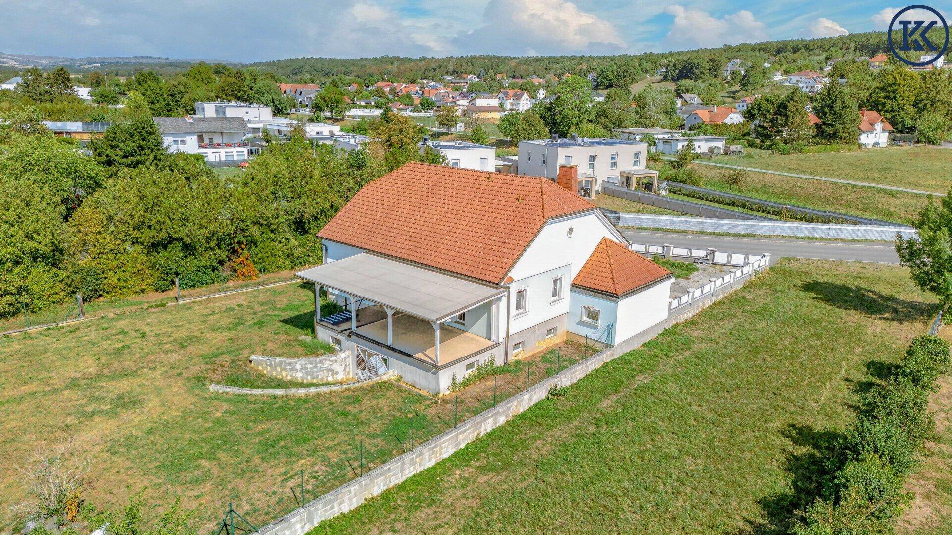 Luftaufnahme des Hauses mit rotem Ziegeldach, heller Fassade und überdachter Veranda, umgeben von Grün.