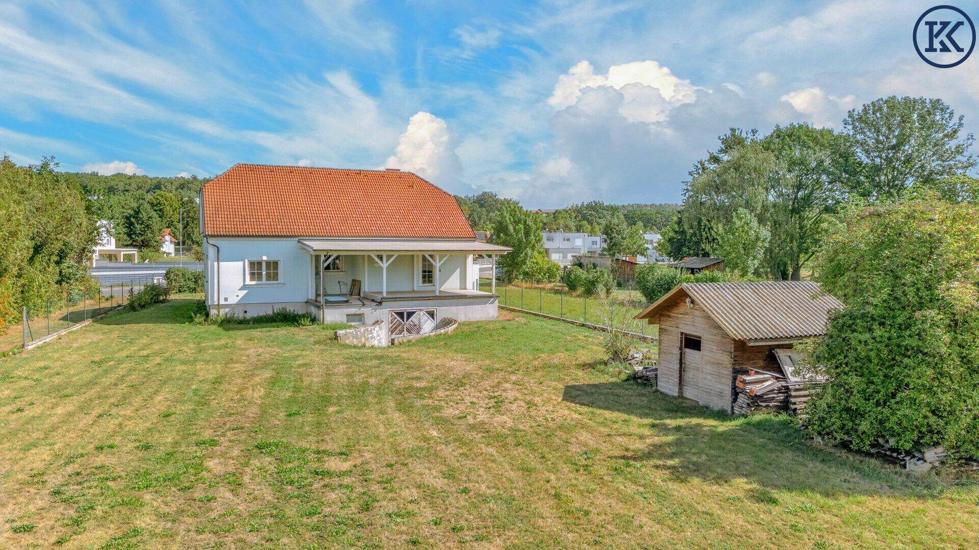 Weitläufiges Gartengrundstück mit Einfamilienhaus, Holzschuppen und viel Grünfläche unter blauem Himmel.