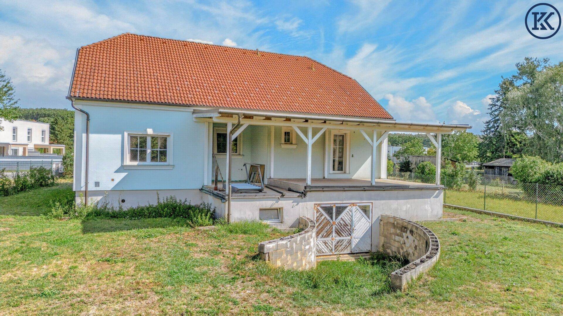 Nahaufnahme des Hauses mit überdachter Veranda und dem geschwungenen Kellerzugang, umgeben von Rasen.