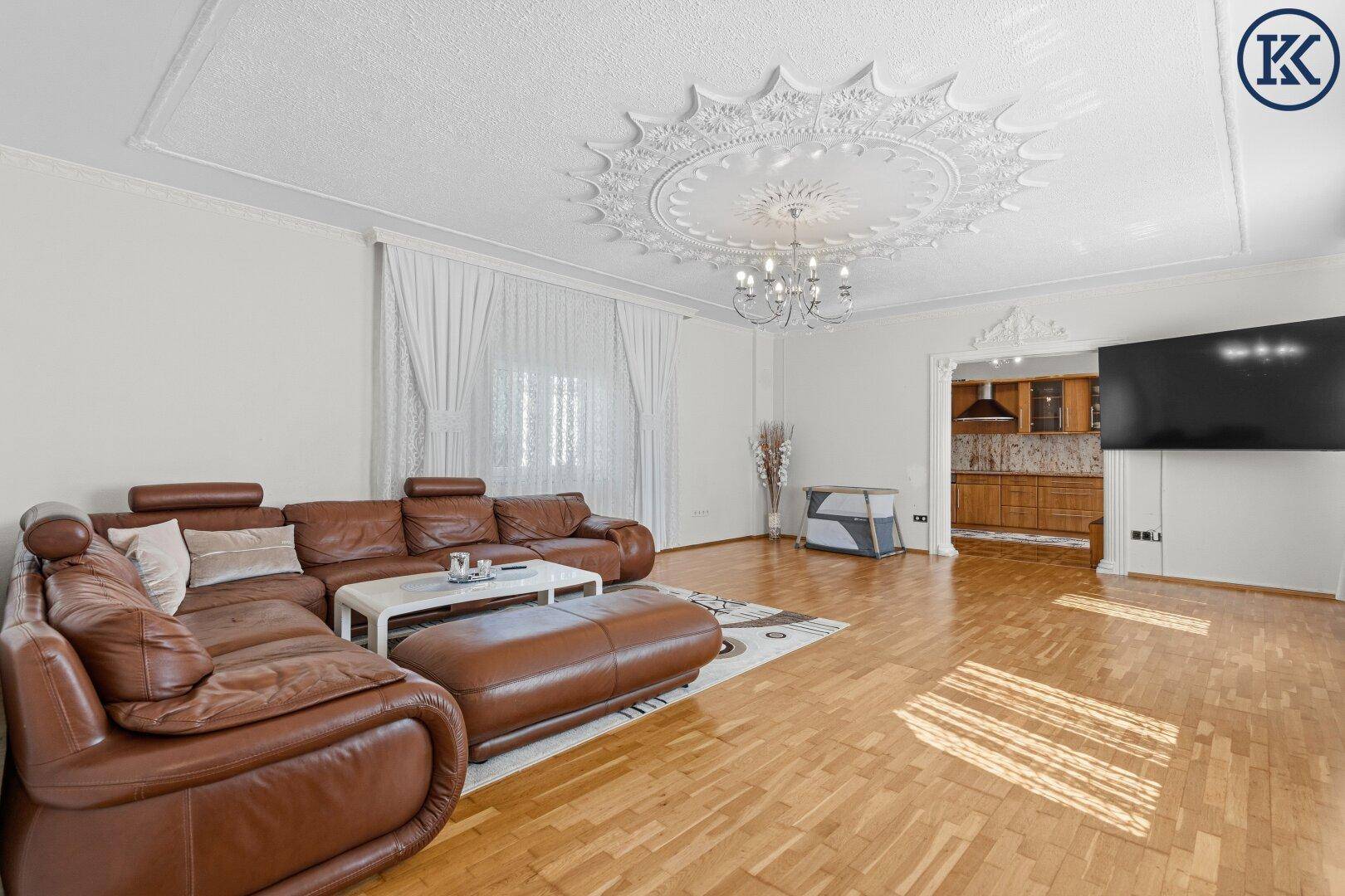 Großes Wohnzimmer mit brauner Ledercouch, Parkettboden und dekorativer Stuckdecke.