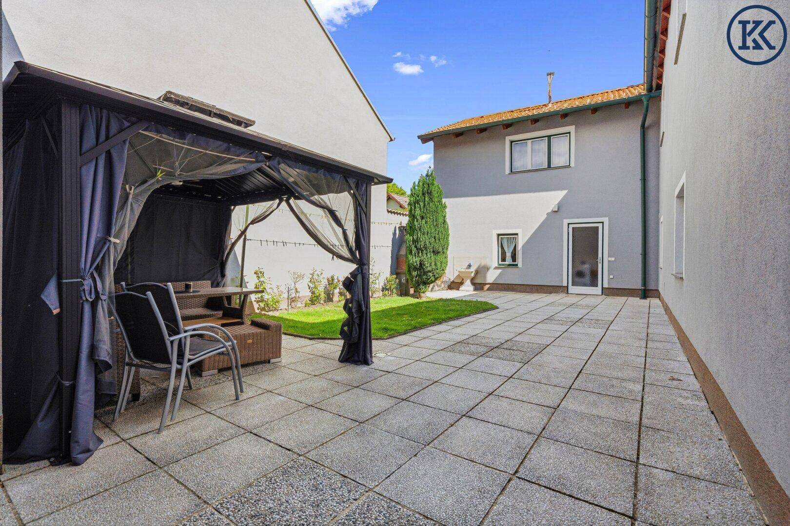 Weitläufige gepflasterte Terrasse mit Gartenlaube und Blick auf die umliegenden Gebäude.