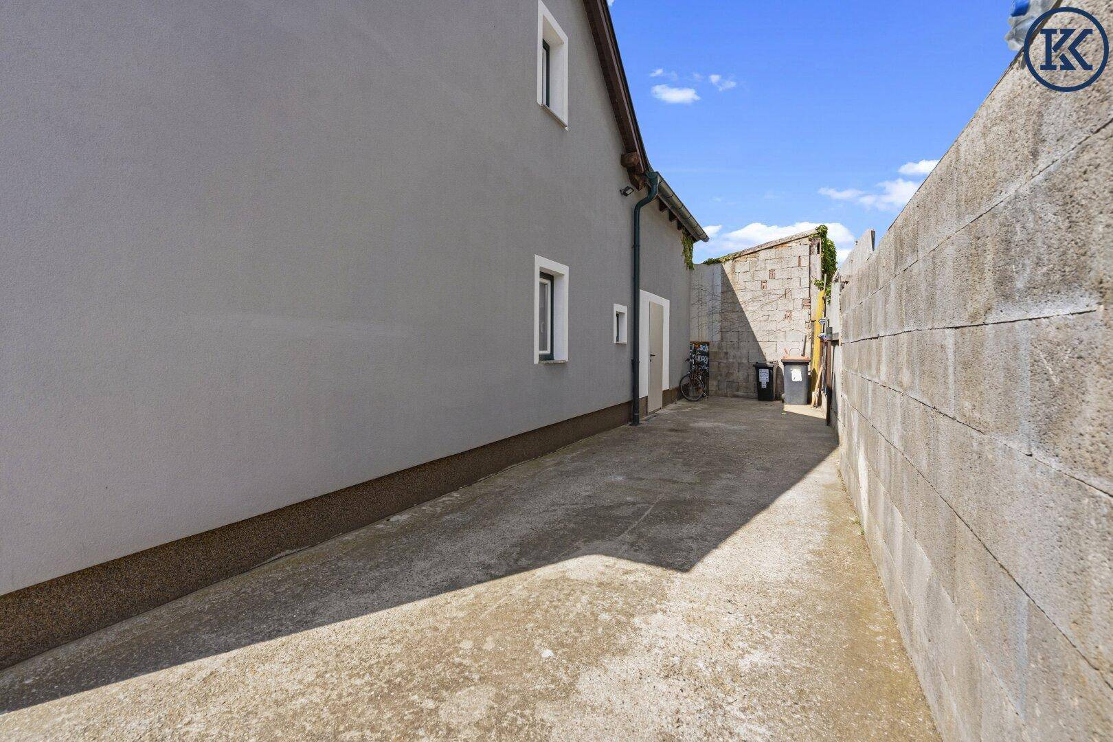 Schmaler gepflasterter Weg entlang des Gebäudes mit hoher Mauer.